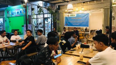 Diskusi publik di kafe dengan peserta duduk, memegang minuman; spanduk biru di latar belakang.