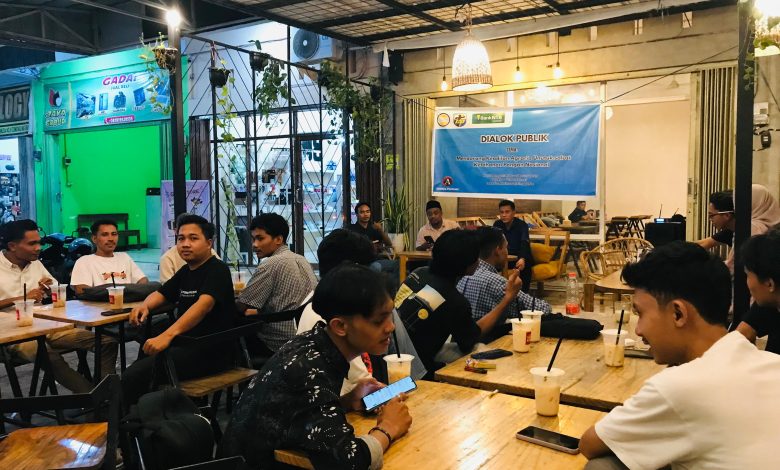 Diskusi publik di kafe dengan peserta duduk, memegang minuman; spanduk biru di latar belakang.