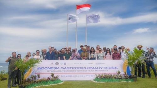 Diplomatic Tour Goes to West Nusa Tenggara." Tiga tiang bendera berdiri di belakang mereka, termasuk satu bendera Indonesia. Acara berlangsung di The Mandalika pada tanggal 8-10 Mei 2023. Lokasinya tampak seperti area terbuka dengan pemandangan laut. (Iam/Ist)